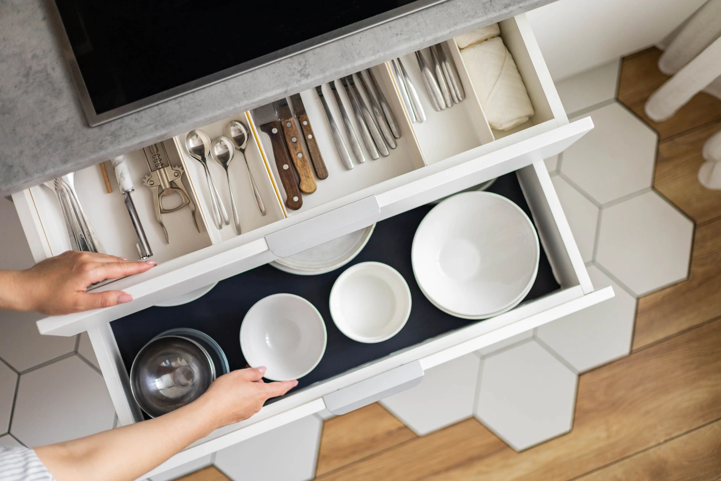 Person opens two white kitchen drawers, one with cutlery, the other with neatly stacked bowls and plates on hexagon tiles.