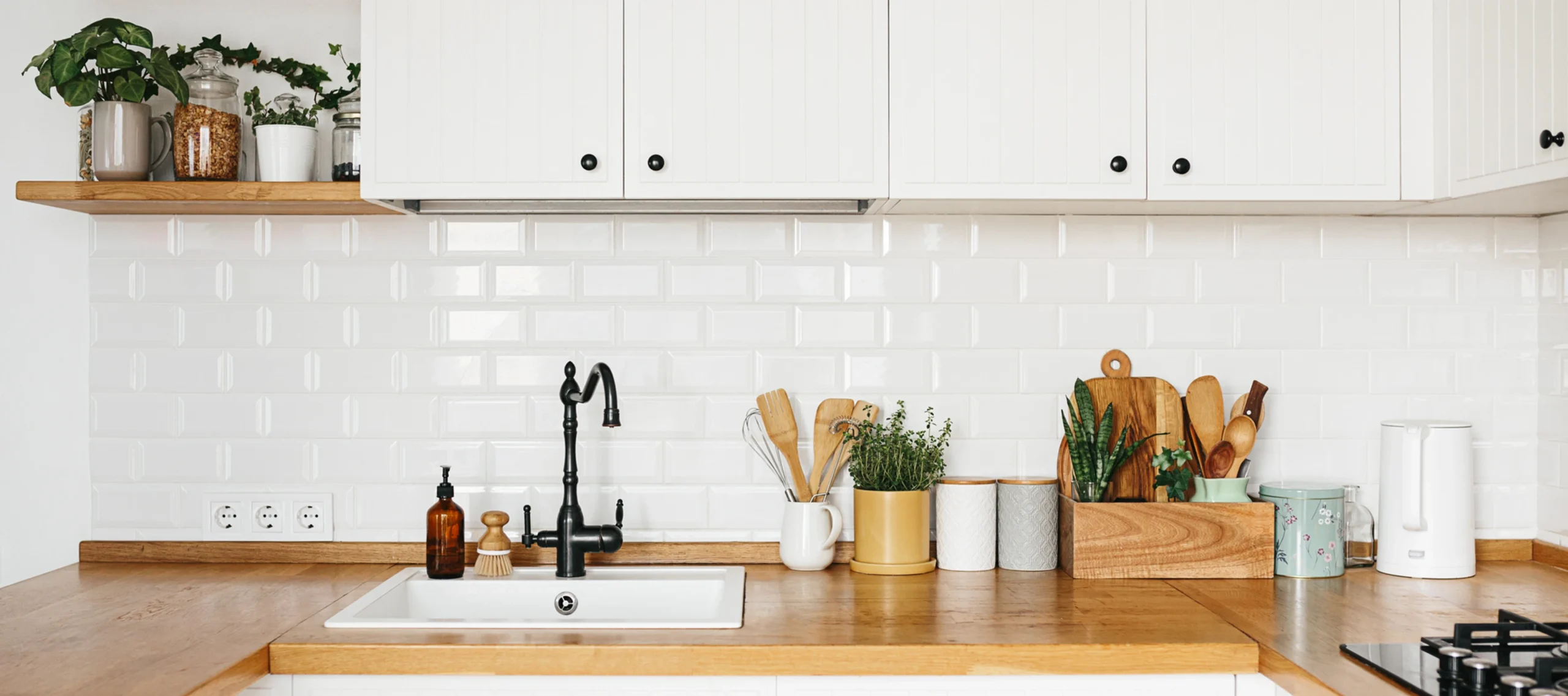 Modern kitchen countertop with white cabinets, black faucet, eco-friendly backsplash, plants, utensils, jars.