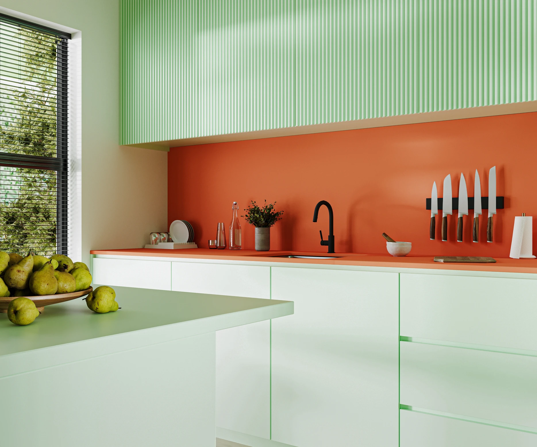 Modern kitchen with mint green cabinets, orange backsplash. Black faucet, knife rack, bowl of pears; sunlight through blinds.