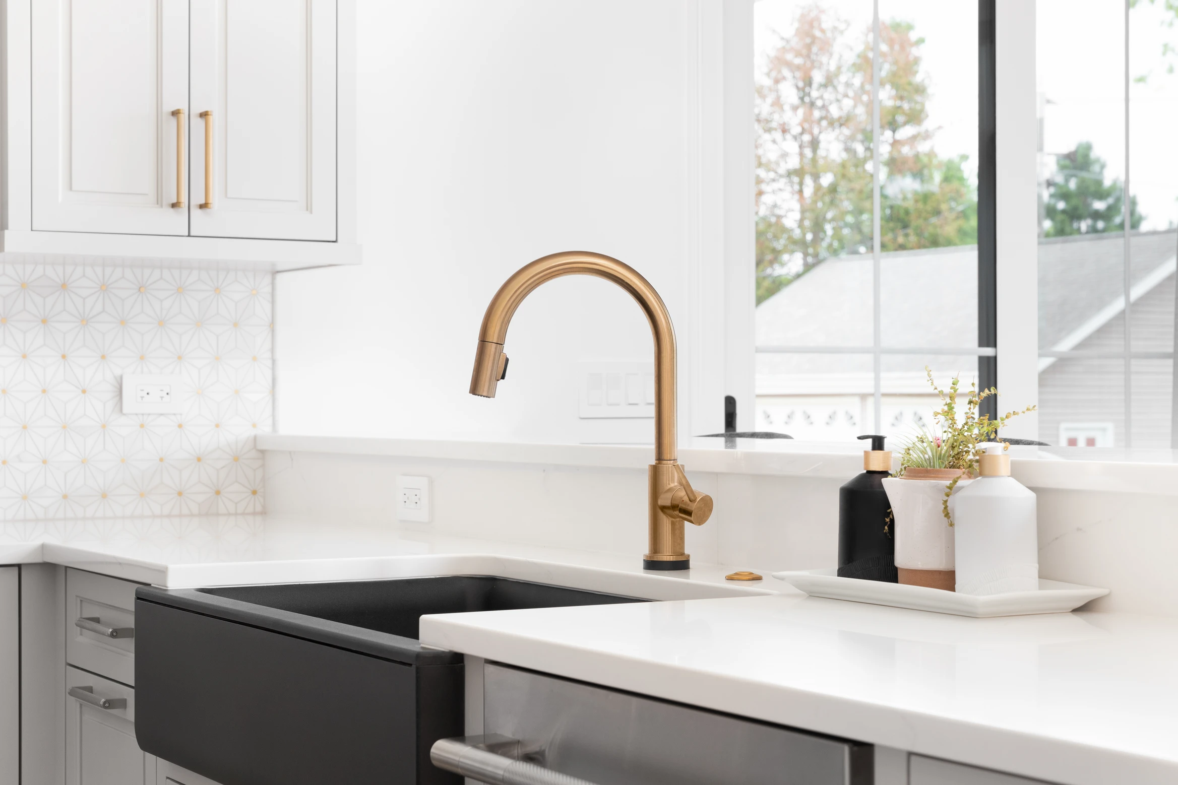 Modern kitchen sink with matte black farmhouse basin, gold faucet, white counters. Soap dispensers near window.