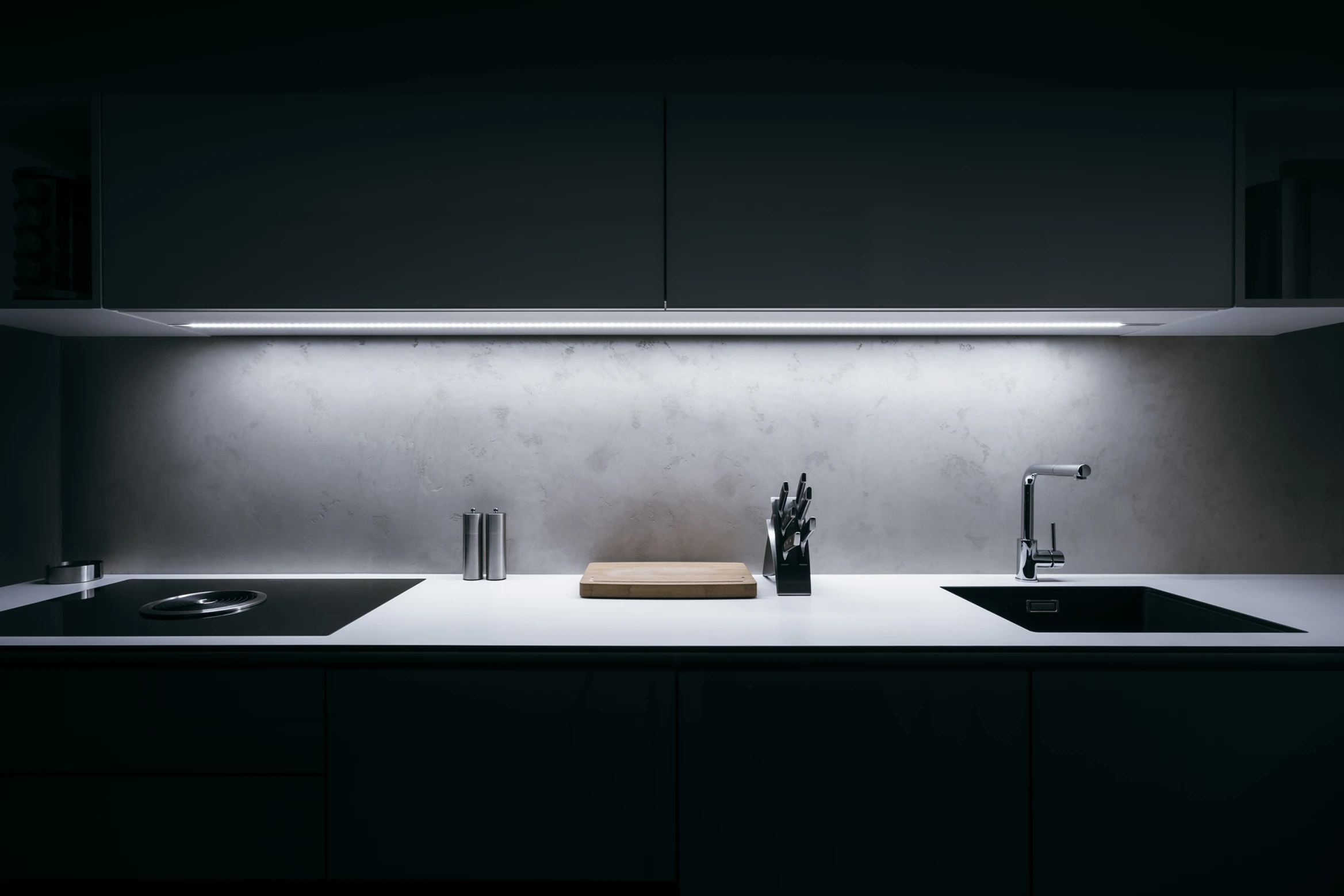 Modern, minimalist kitchen with white countertop, cutting board, knife block, stovetop and statement lighting.