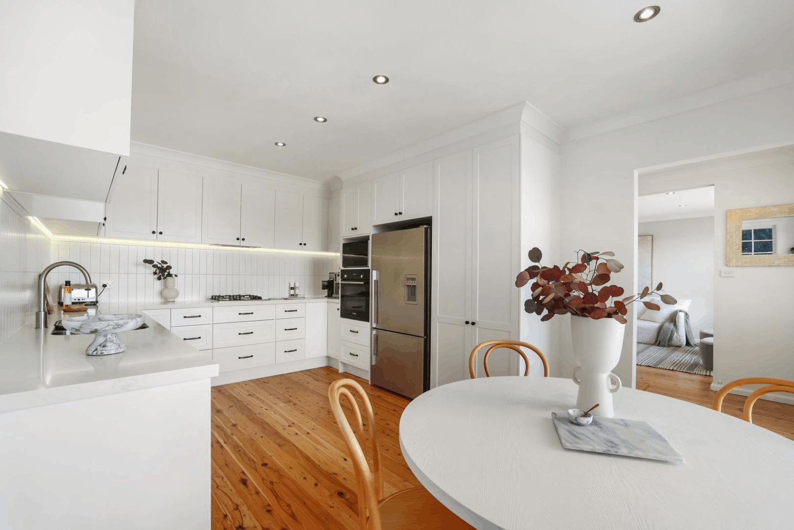 Modern white kitchen with wooden floors, round table, light chairs, stainless steel appliances—Red Hot Kitchens style.