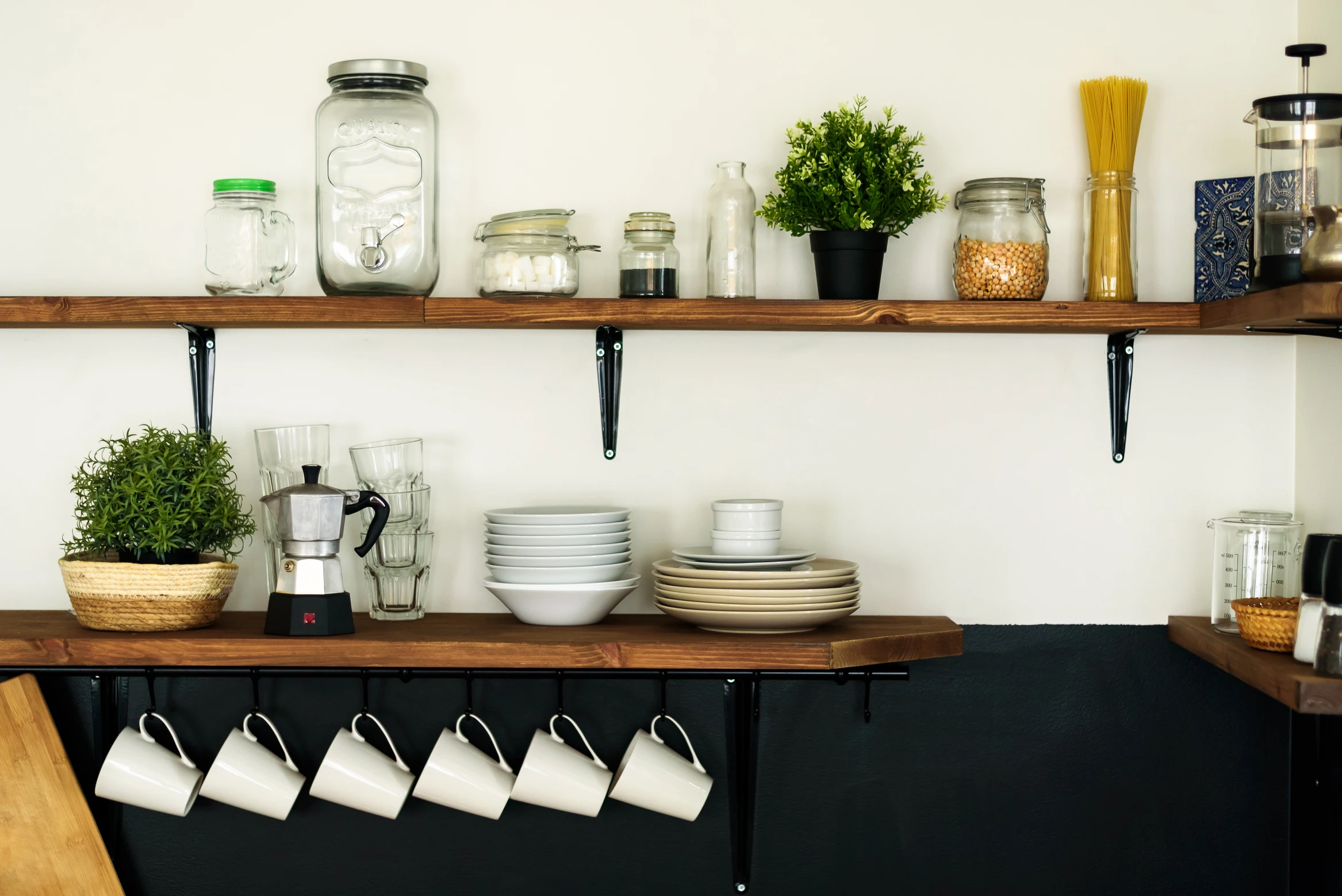 Two wooden open shelving units showcase jars, glassware, plates, bowls, mugs, coffee maker & small plants by white wall.