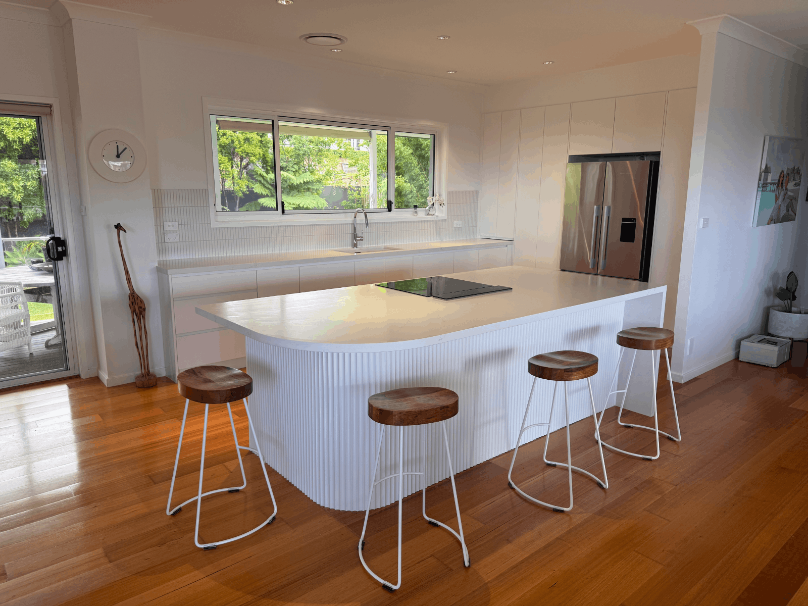 Modern kitchen makeover by Red Hot Kitchens: white island, cooktop, wood stools, stainless fridge, large window.