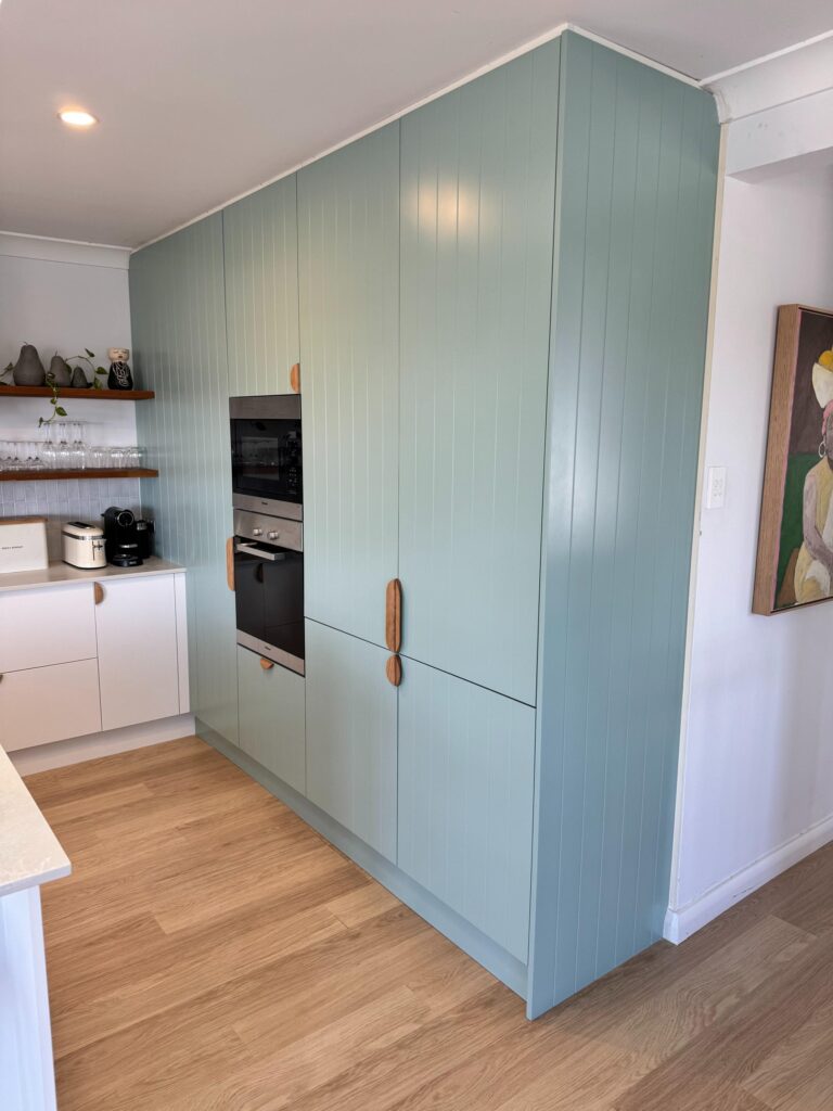 Modern kitchen on Collaroy Plateau with blue-green cabinets, wood handles, white countertops, and open shelves.