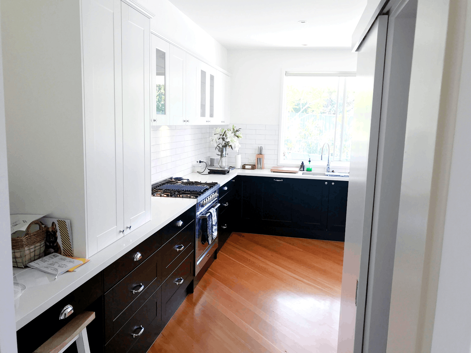 Modern kitchen with white uppers, black lowers, stainless appliances, wood floor & bright window. Red Hot Kitchens.