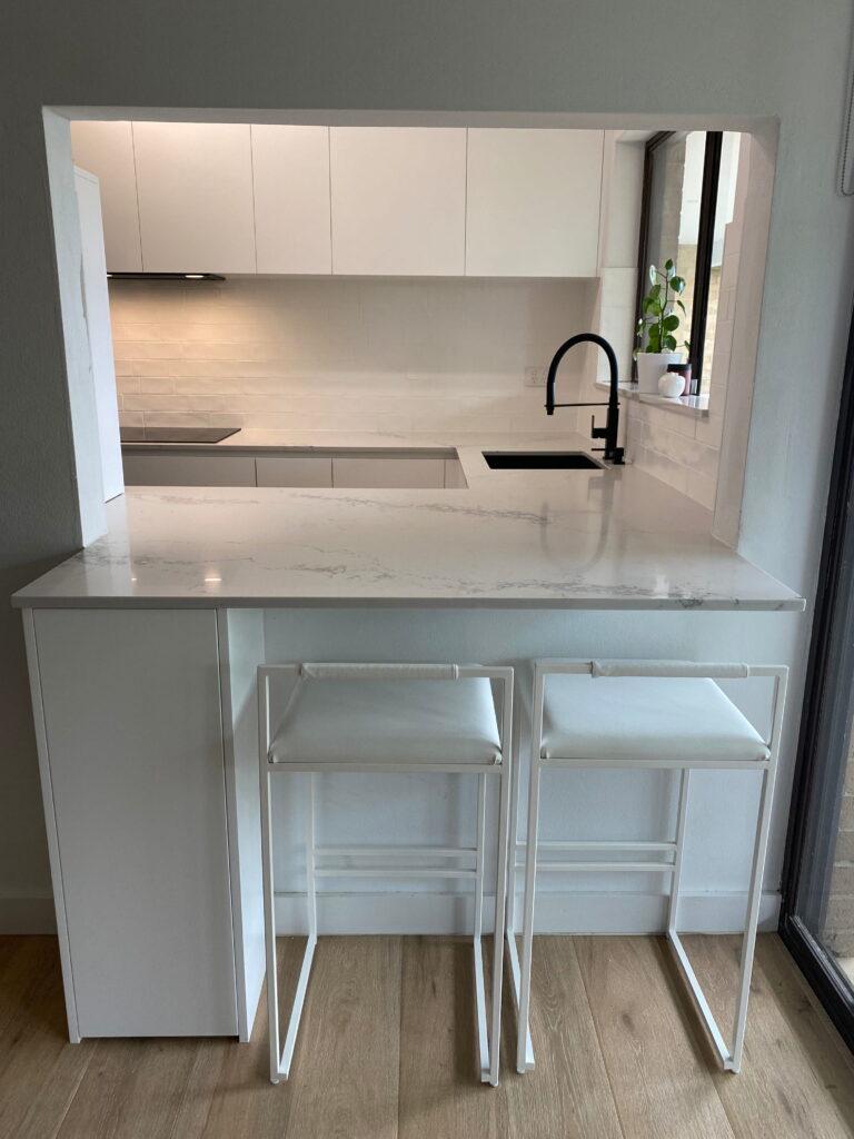 White kitchen with a marble countertop breakfast bar and two white metal bar stools facing a window.