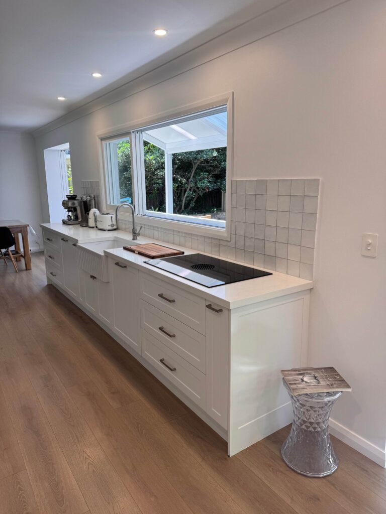 Red Hot Kitchens modern white kitchen in Mona Vale, wood floors, big windows, sleek counter & metallic stool.