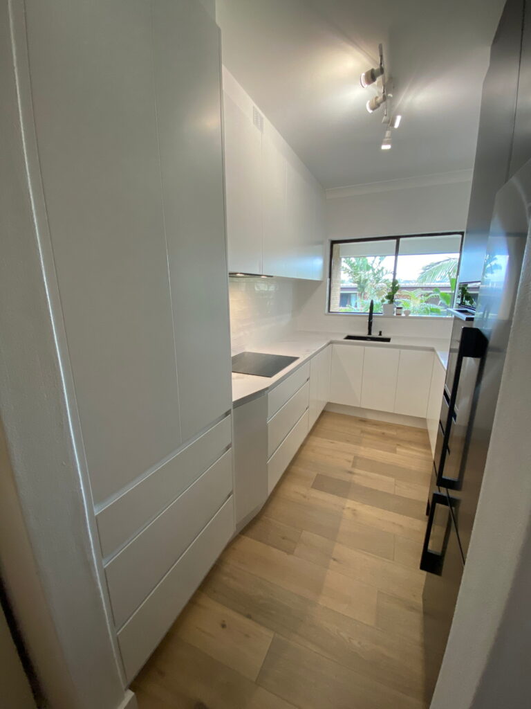 Modern galley kitchen by Red Hot Kitchens with white cabinets, wood floor, big window over sink, sleek track lights