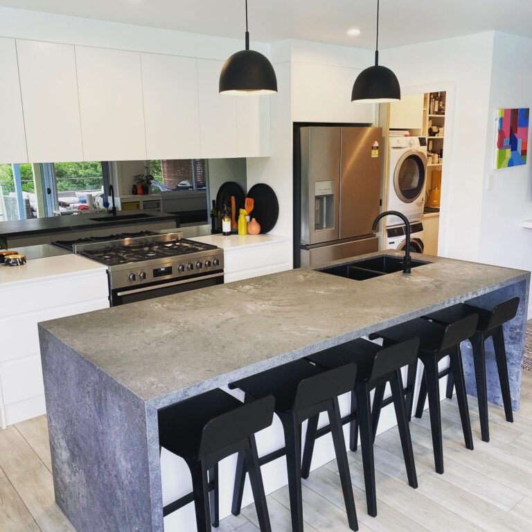 Modern white kitchen with a large grey stone island and black bar stools along the island. Stainless steel refrigerator and built-in oven visible, plus a laundry area with a washer/dryer in the back. Pendant lights hang from above.