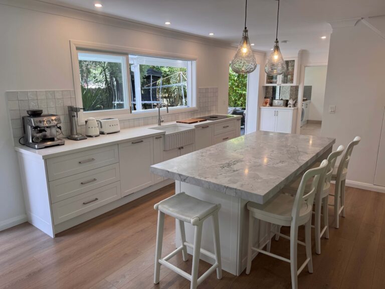 Red Hot Kitchens modern white kitchen with marble island, three chairs, pendant lights, and garden views.
