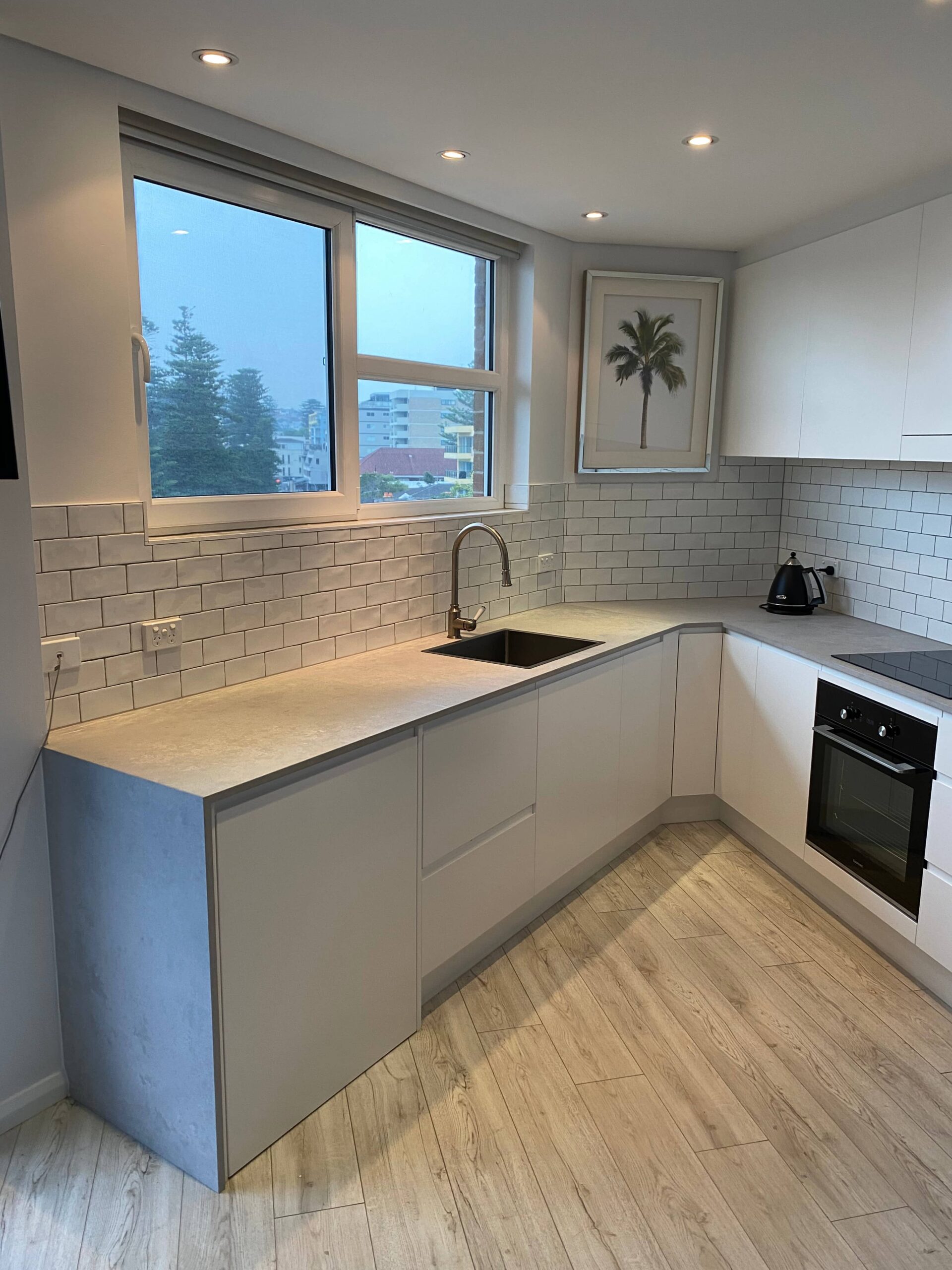 Modern apartment kitchen with white cabinets, light wood floors, built-in oven, and palm tree artwork.