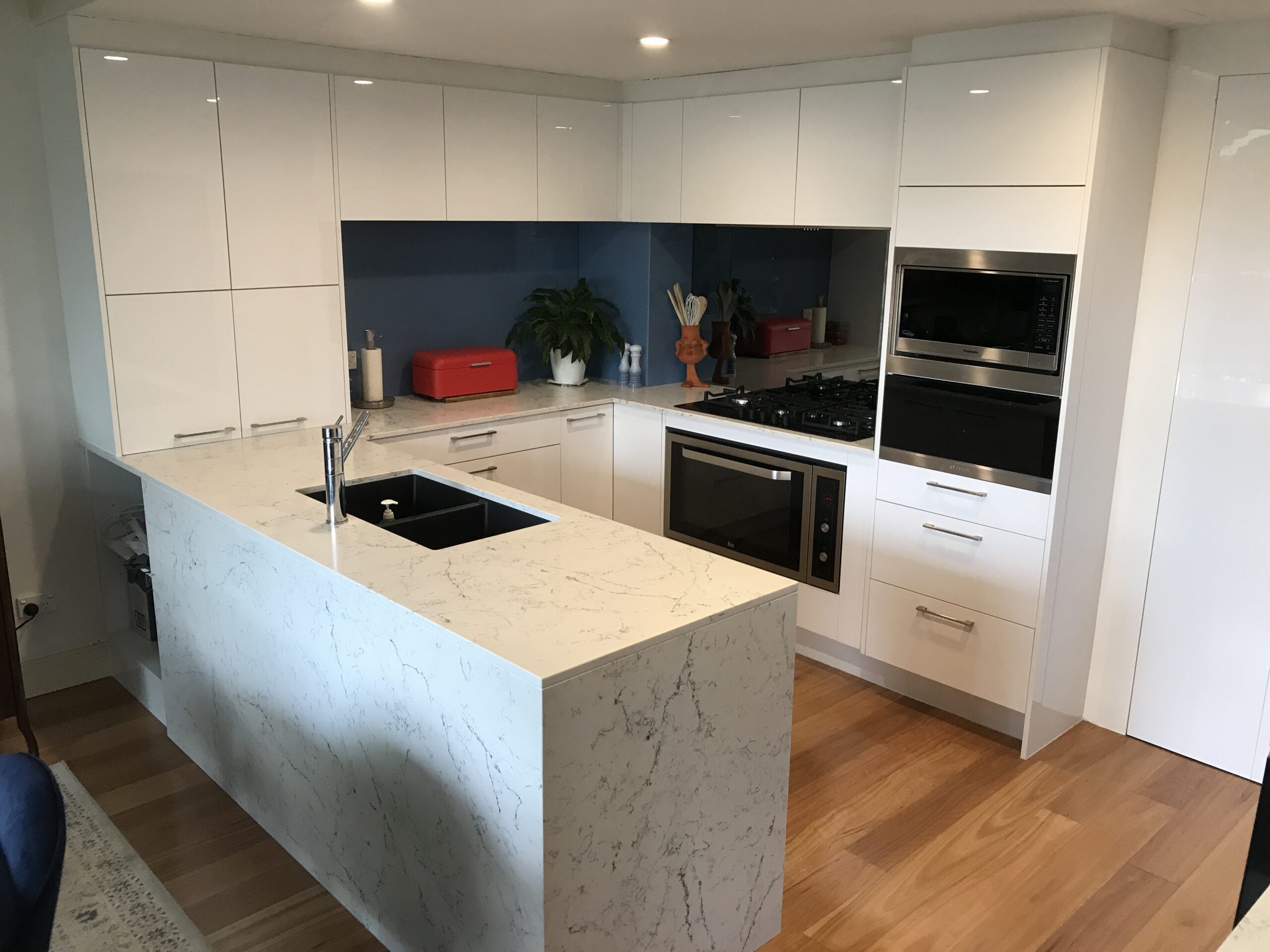 Modern kitchen by Red Hot Kitchens in Brookvale with white cabinets, marble island, red toaster, and wood floor.
