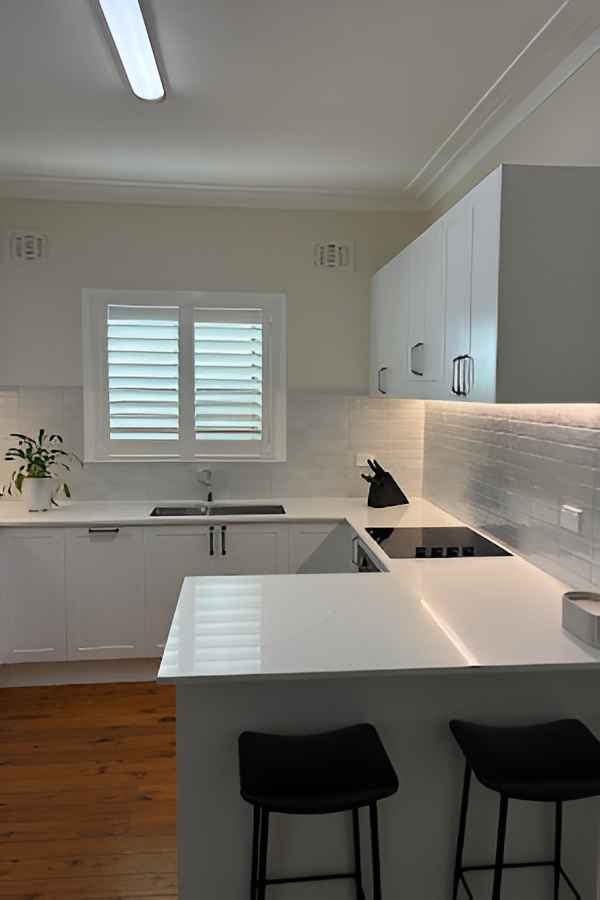 Modern white kitchen with custom cabinetry, black stools, peninsula, plant, and window by Red Hot Kitchens.