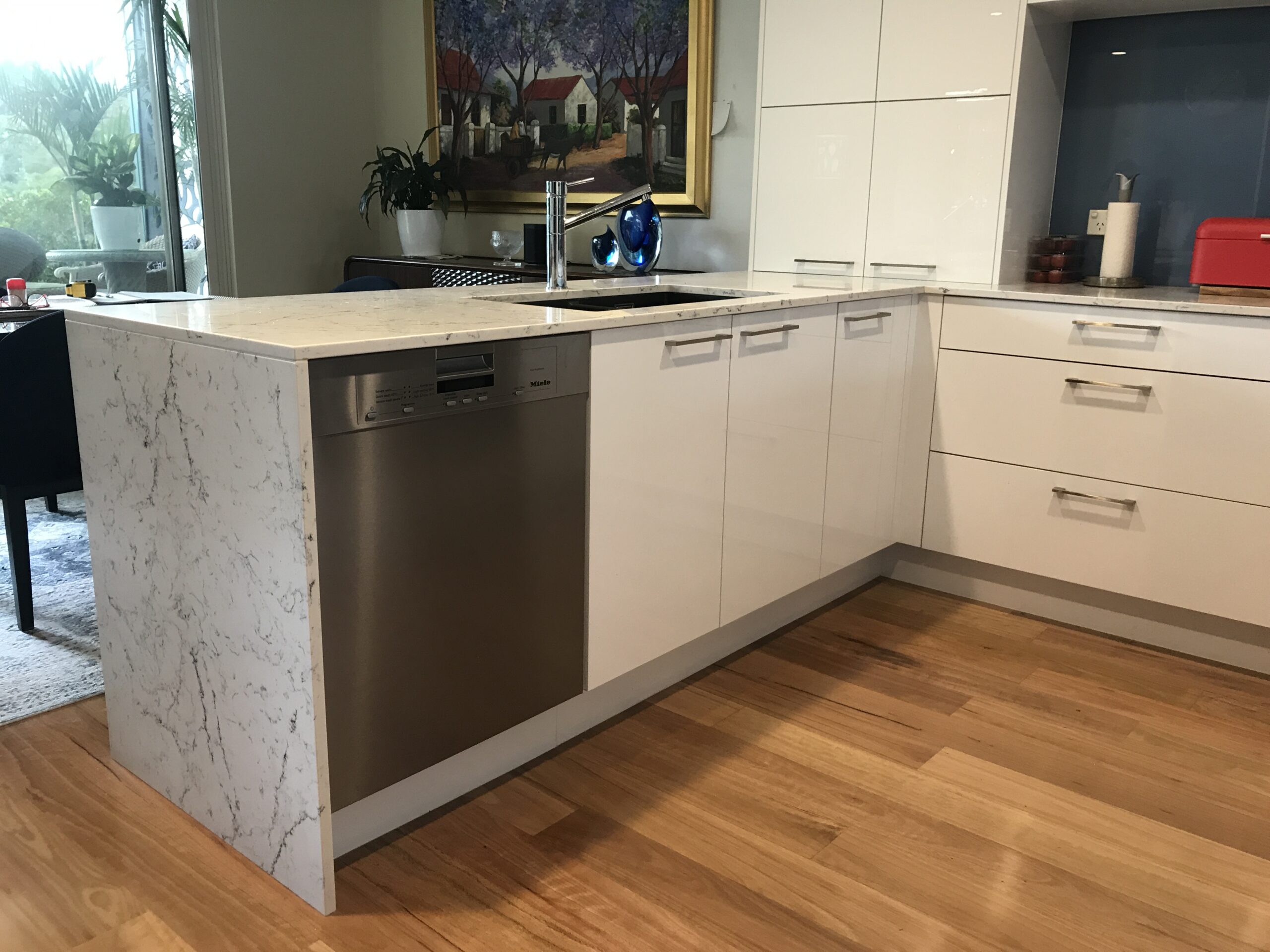 Modern kitchen with white cabinets, stainless steel dishwasher, marble countertop, wood floor by Red Hot Kitchens Brookvale.