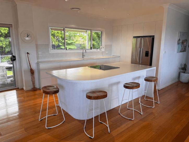 Modern kitchen with white cabinets, island, wood stools and floors by Red Hot Kitchens; window to green view.