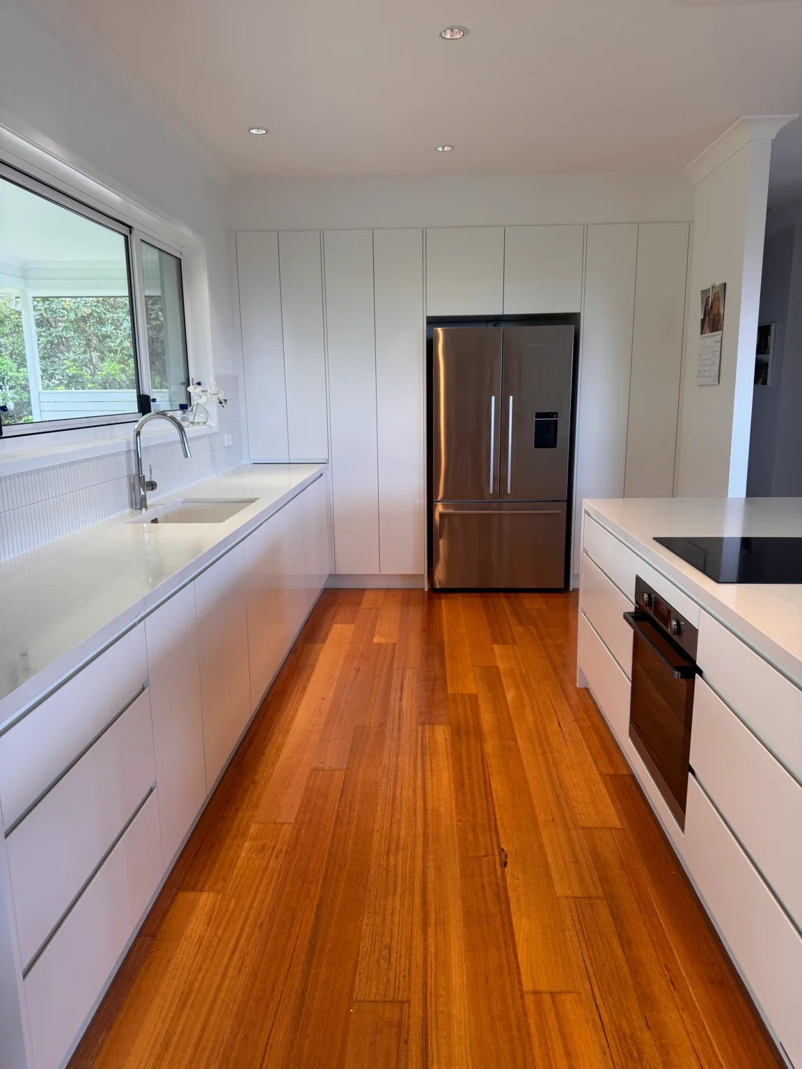 White handleless kitchen cabinetry beneath a wide window, with tall cupboards surrounding a stainless-steel fridge in Allambie Heights.