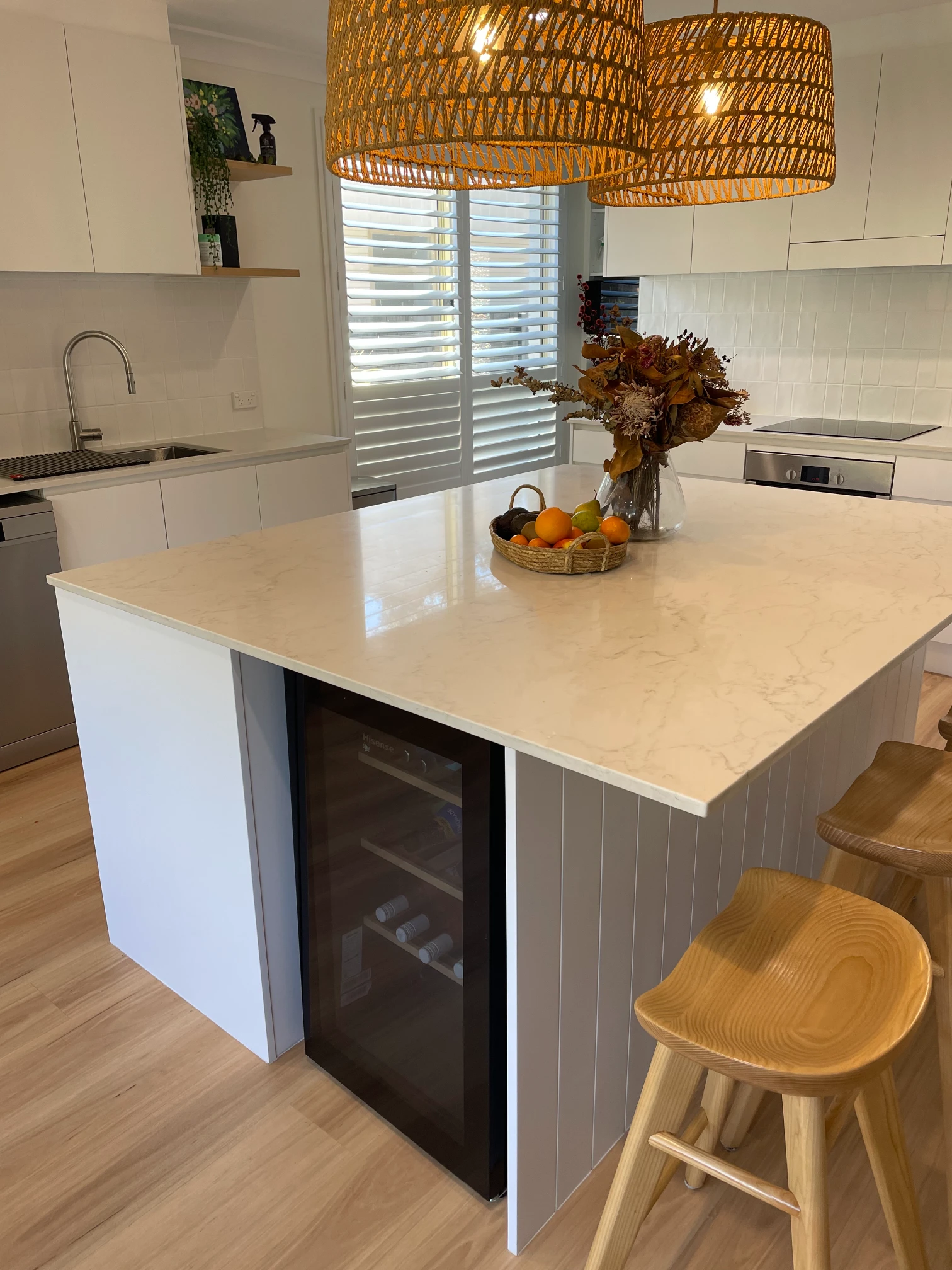 Cromer kitchen island with a built-in wine fridge, pale benchtop and timber stools beneath woven pendant lights.