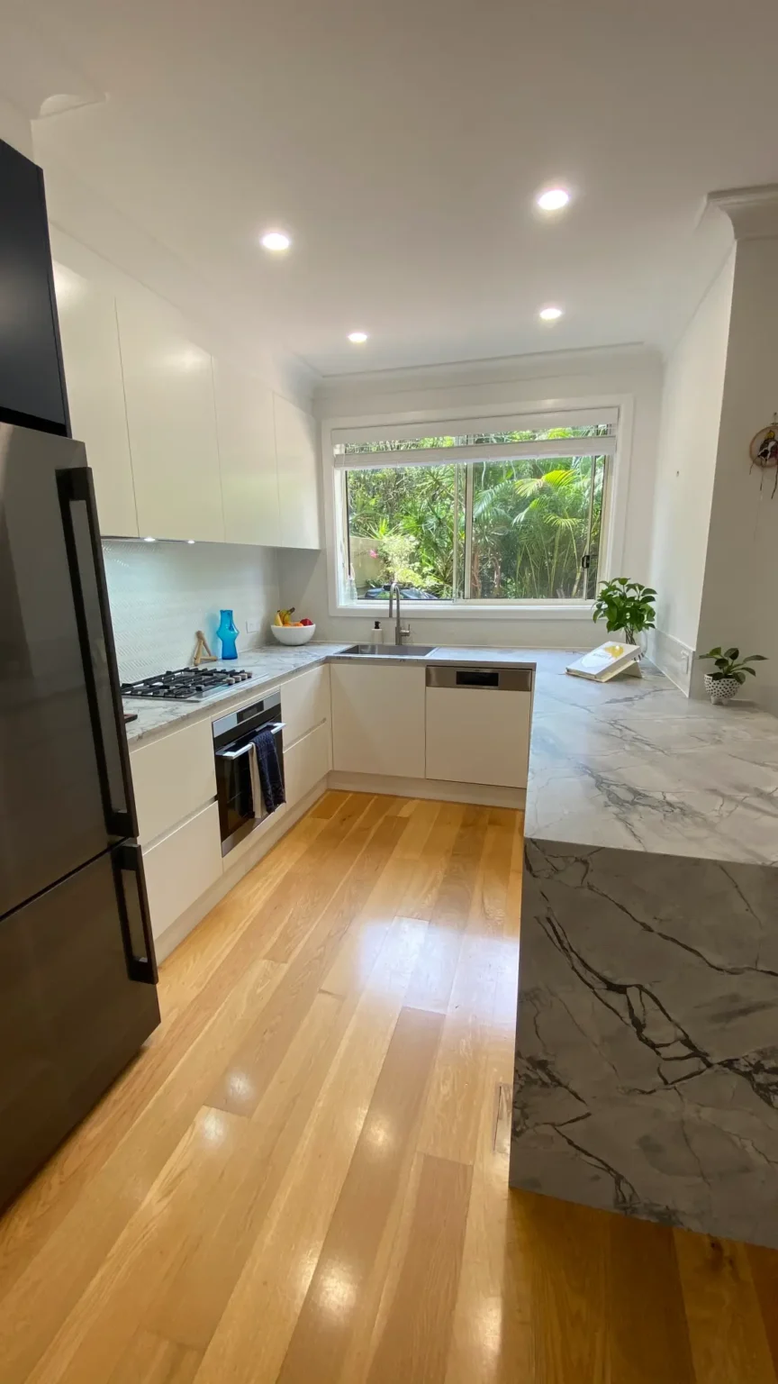 Modern kitchen renovation in Dee Why featuring white cabinets, a marble benchtop, wooden floors, and a window view.