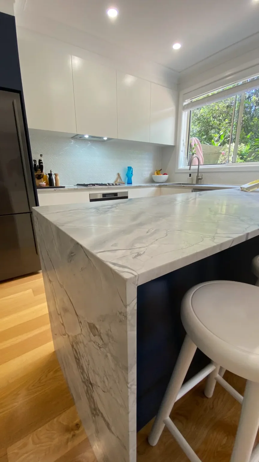 Modern kitchen in Dee Why featuring a marble waterfall island bench, white cabinets, and wooden floors in this renovation.