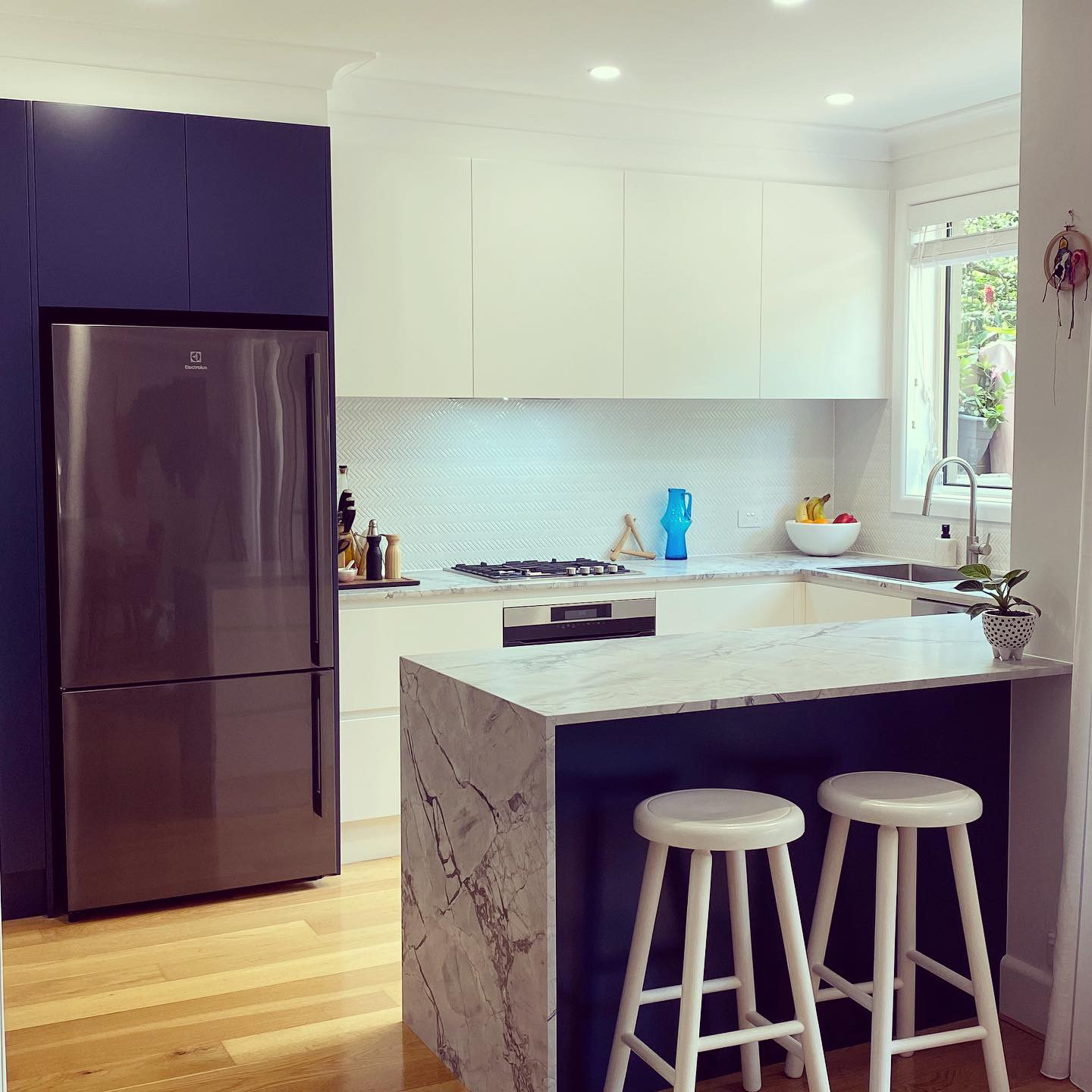 Modern kitchen renovations in Dee Why with a stone island bench, two white stools, and dark blue cabinetry.