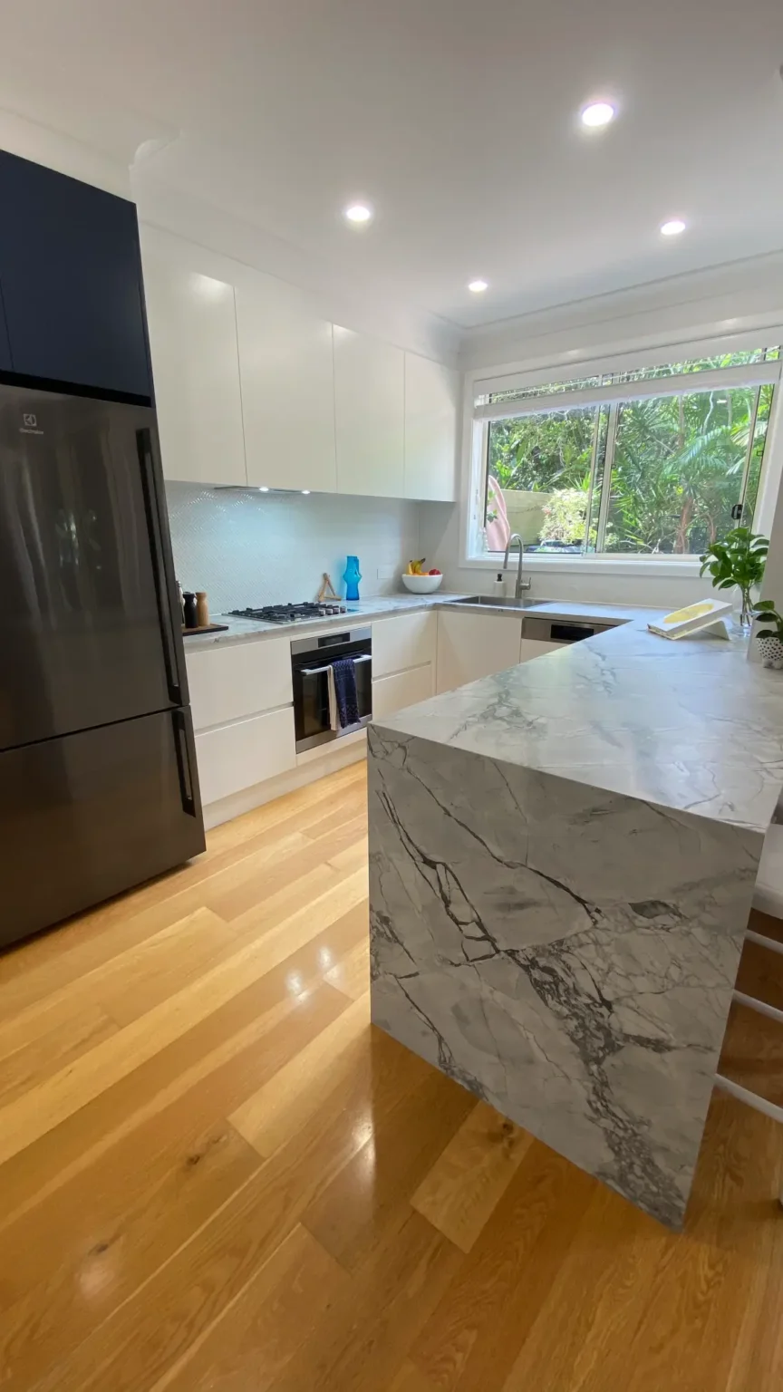 Modern kitchen renovation featuring a white marble island, light wood flooring, and black appliances, white cabinetry.
