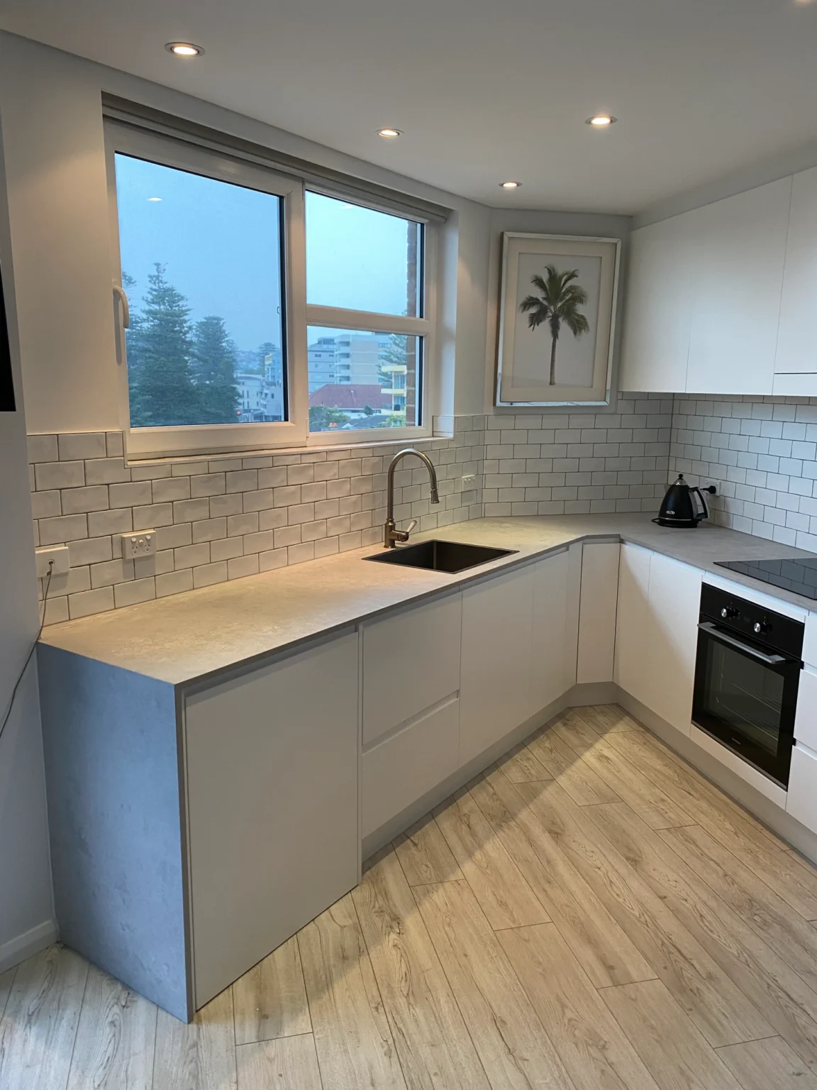 Modern kitchen renovation with white cabinets and black sink, featuring a view of Manly through the window.