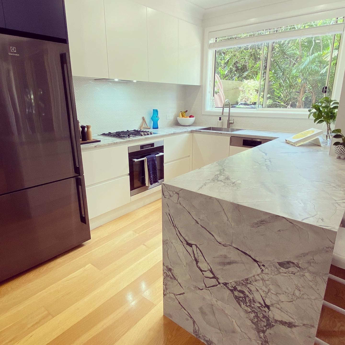 Modern white kitchen renovation featuring a large marble island, sleek cabinets, and a dark stainless refrigerator.