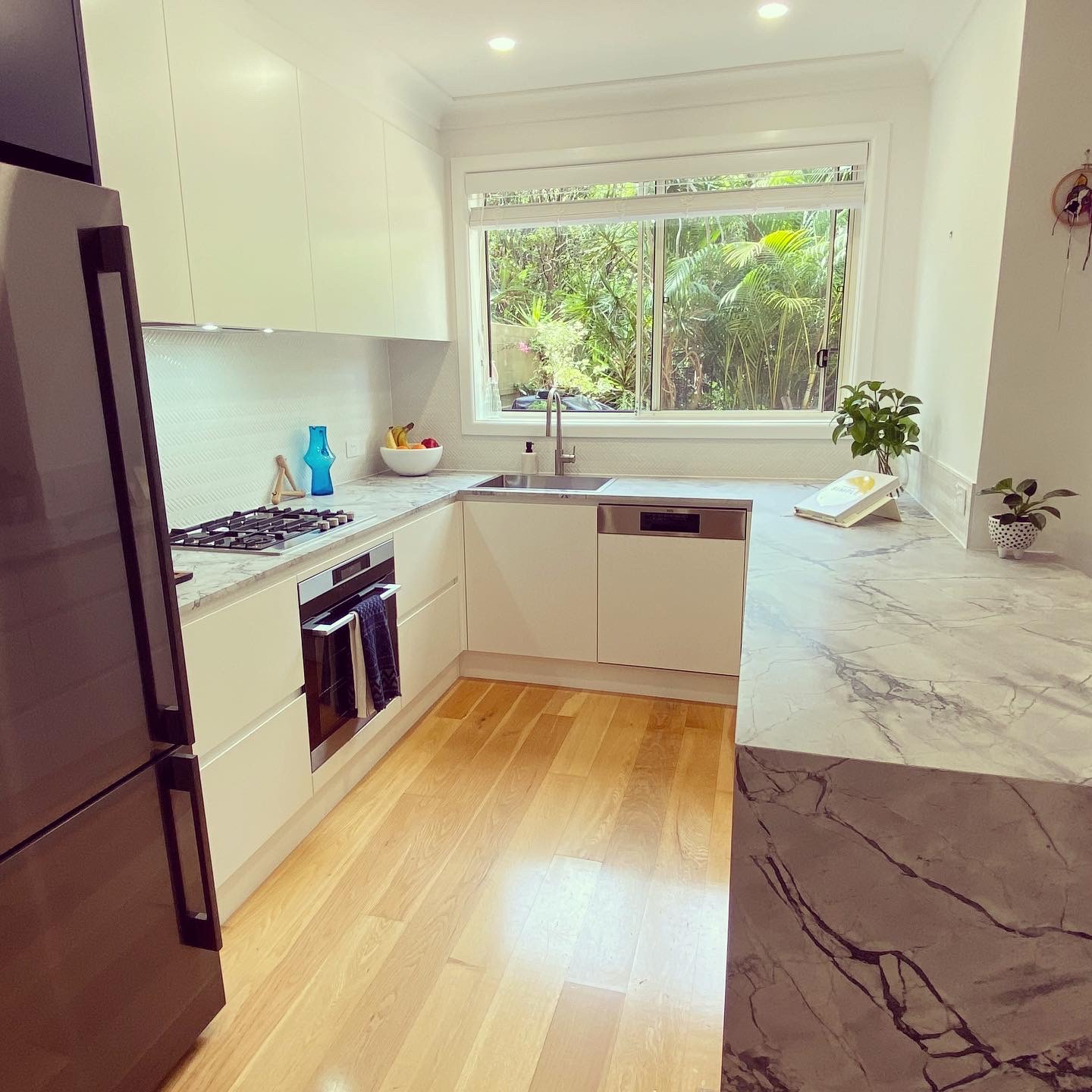 Modern white U-shaped kitchen with marble countertops, light wood flooring, and garden views, a Dee Why renovation.